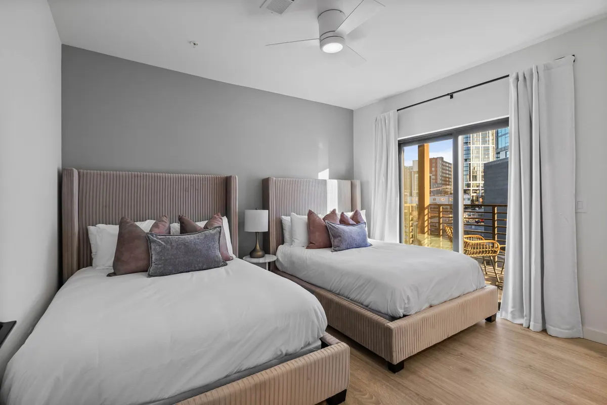 A bright secondary bedroom with two queen beds featuring plush headboards and decorative pillows. A large window offers a view outside, and the space feels inviting and perfect for groups or families.