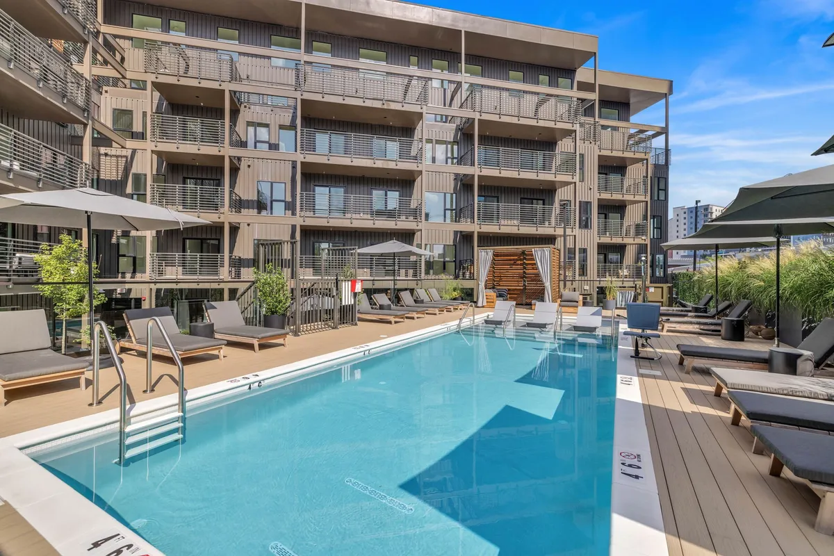 A serene outdoor swimming pool surrounded by lounge chairs and greenery, with the building facade in the background. The pool area looks inviting for both swimming and relaxing, with ample space for social gatherings and sunbathing.