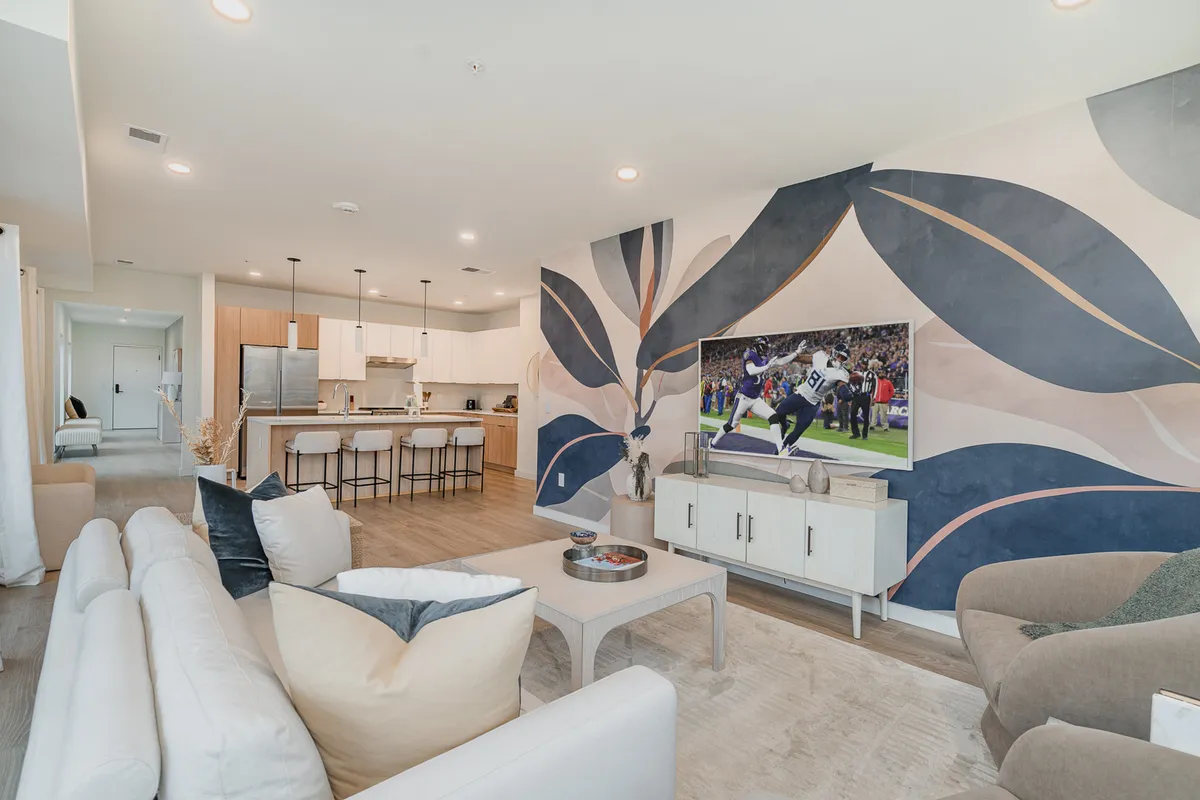 The image showcases a stylish and modern open-concept living area with a vibrant tropical wall mural, plush sofa arrangement facing a large television, and a sleek kitchen area in the background. The scene is set for group gatherings, whether for casual dining, watching sports, or simply relaxing with friends and family.