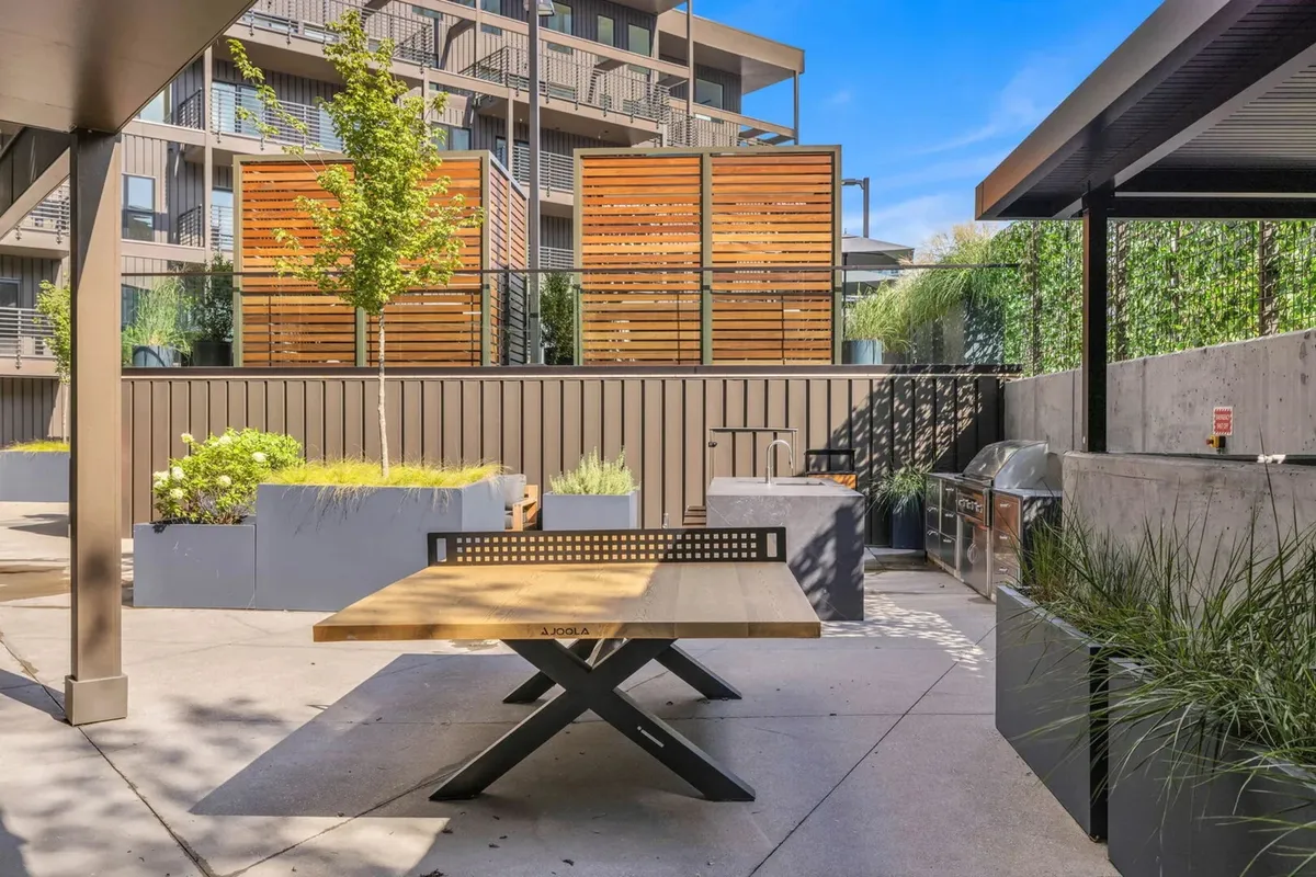 An exterior community BBQ area is captured here with a wooden table, planters, privacy screens, and a built-in outdoor grill. The setting is casual and communal, ideal for group meals or evening gatherings under the stars.