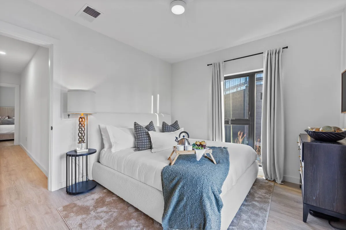 A serene bedroom featuring a king-sized bed adorned with decorative pillows and a cozy throw blanket. Natural light streams in through a window, and a nightstand with a lamp and a dresser add to the inviting, restful atmosphere.