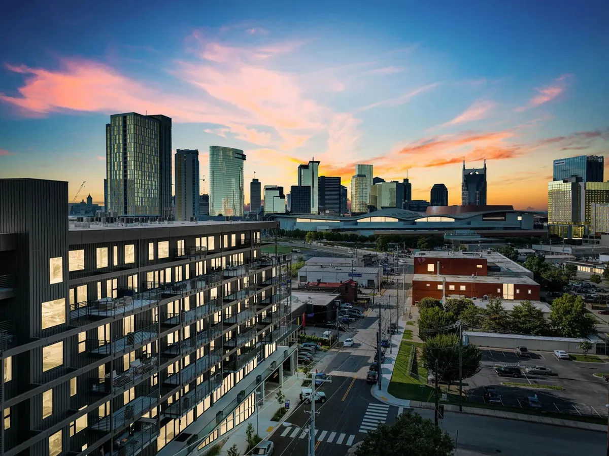 A striking view of a modern apartment building set against the Nashville city skyline at sunset. The image captures the vibrancy of the urban setting, with a focus on the building’s contemporary design and the lively, colorful sky that hints at the city’s dynamic atmosphere and nightlife.