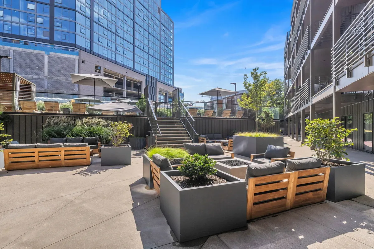 A modern outdoor communal space featuring stylish seating areas, planters with greenery, sun loungers, and a canopy-covered area. The setting is tranquil and designed for both relaxation and socializing, with hints of sunset lighting.
