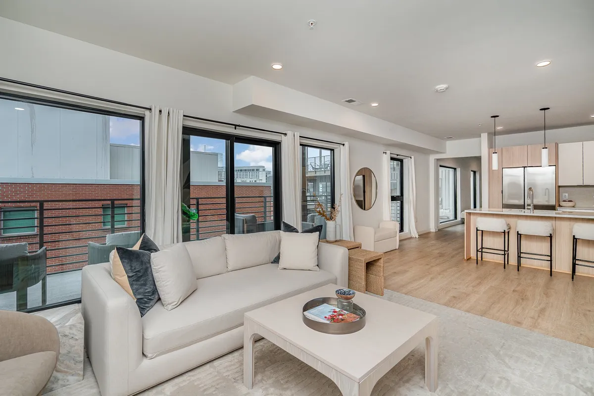 A wide-angle view of the open-concept living area and kitchen, featuring a comfortable sofa, a sleek kitchen island, large windows flooding the space with natural light, and a decorative mirror. The setting is styled for both relaxation and entertaining, with contemporary furnishings and inviting decor.