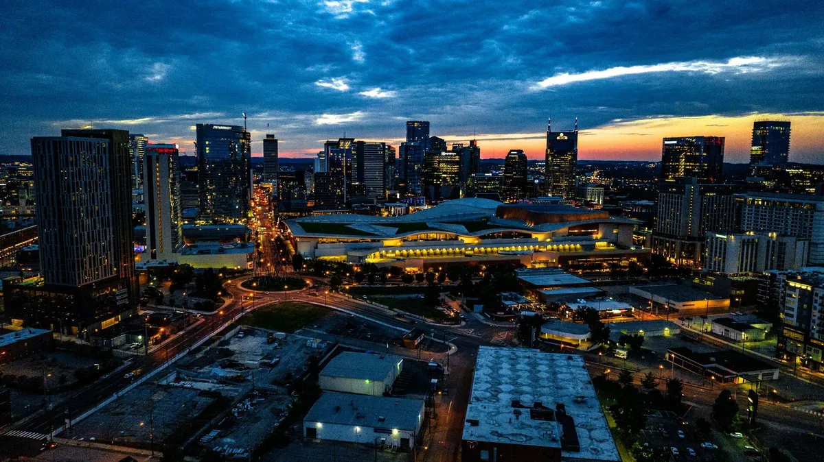 An exterior cityscape at dusk, showing Nashville's skyline with illuminated buildings, street lights, and sunset colors. The energetic mood hints at the bustling nightlife, with an inviting view of the city as it comes alive after dark.