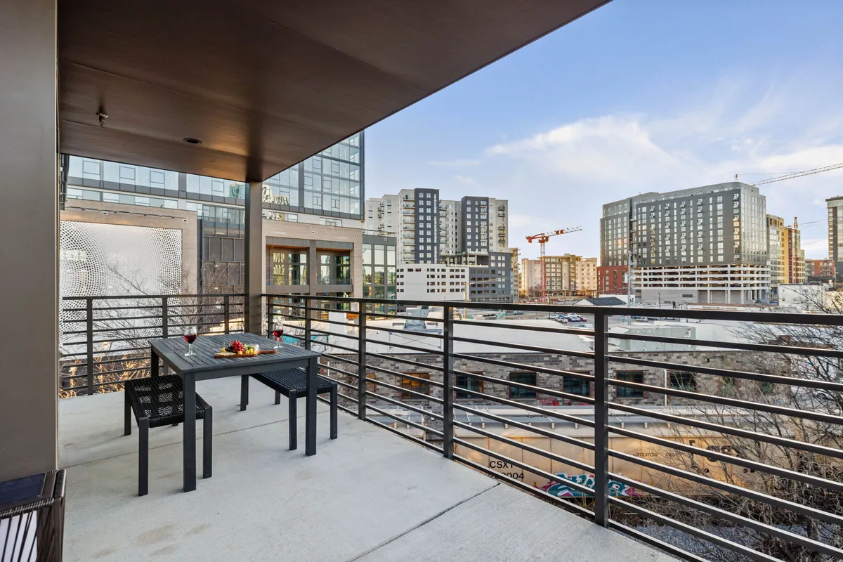 A stylish balcony featuring a cozy seating arrangement and decorative touches, with a stunning view of the city skyline. The table is set for outdoor dining or drinks, and the golden hour lighting enhances the inviting and celebratory mood, ideal for gathering with friends or enjoying a meal al fresco.