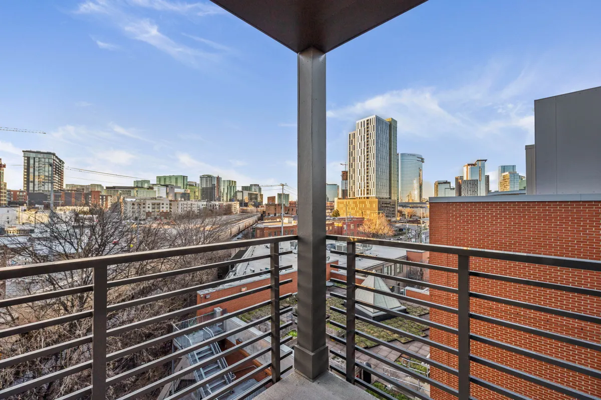 A contemporary balcony scene featuring a stylish railing, views of the city skyline, modern architecture, and nearby trees. The angle suggests a resident enjoying coffee while taking in the urban landscape, hinting at both relaxation and connectivity to the energy of the city below.